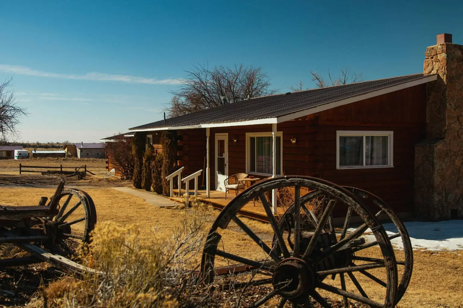A classic log cabin with stone chimney on open rural land, featuring antique wooden wagon wheels in the foreground.